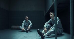 Two men in prison uniforms sitting in a cell, reflecting on their situation under dim lighting.